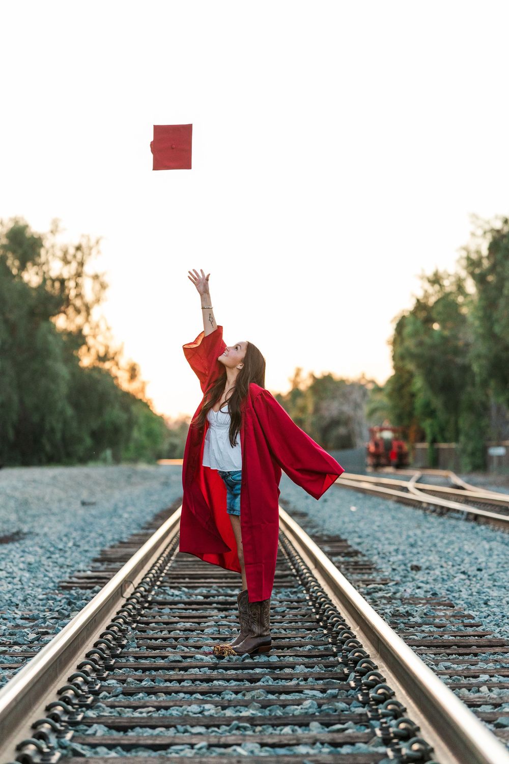 Four sequential photos show a figure in a red graduation gown dancing joyfully along train tracks at sunset.