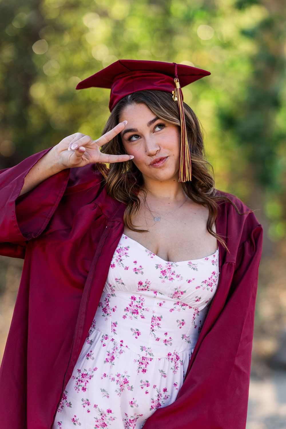 Model wearing maroon varsity jacket and floral dress poses playfully with baseball cap in outdoor portrait session.