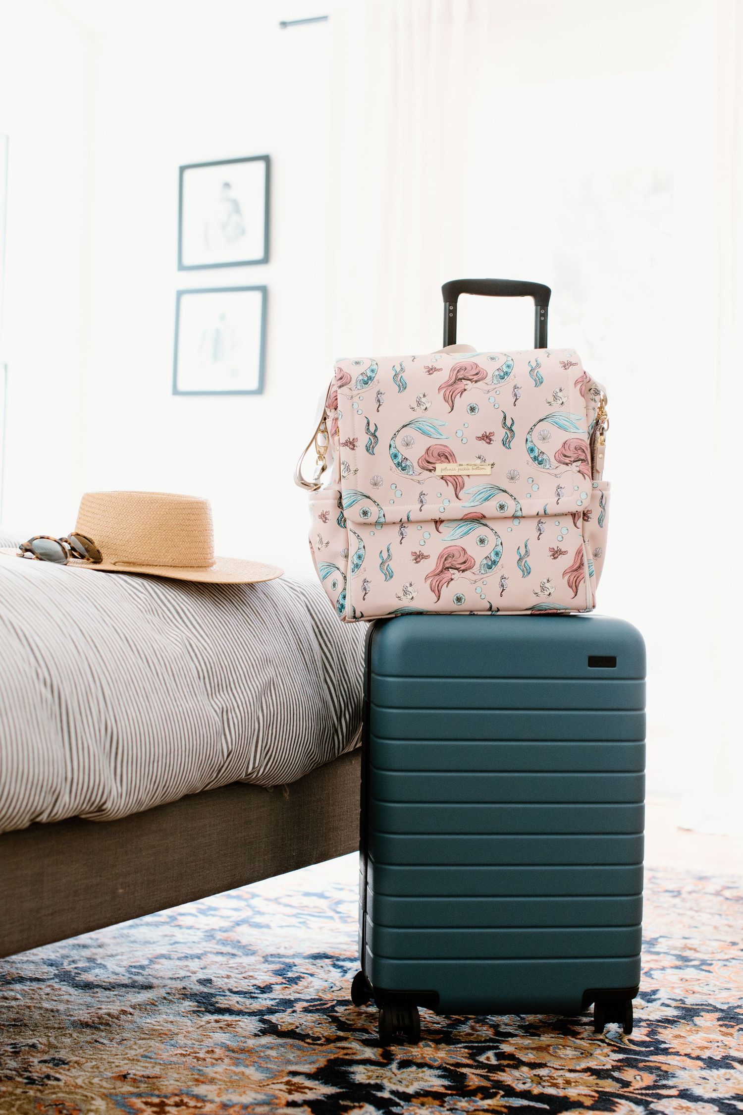 Teal suitcase and pink floral travel bag on bed with beige hat.
