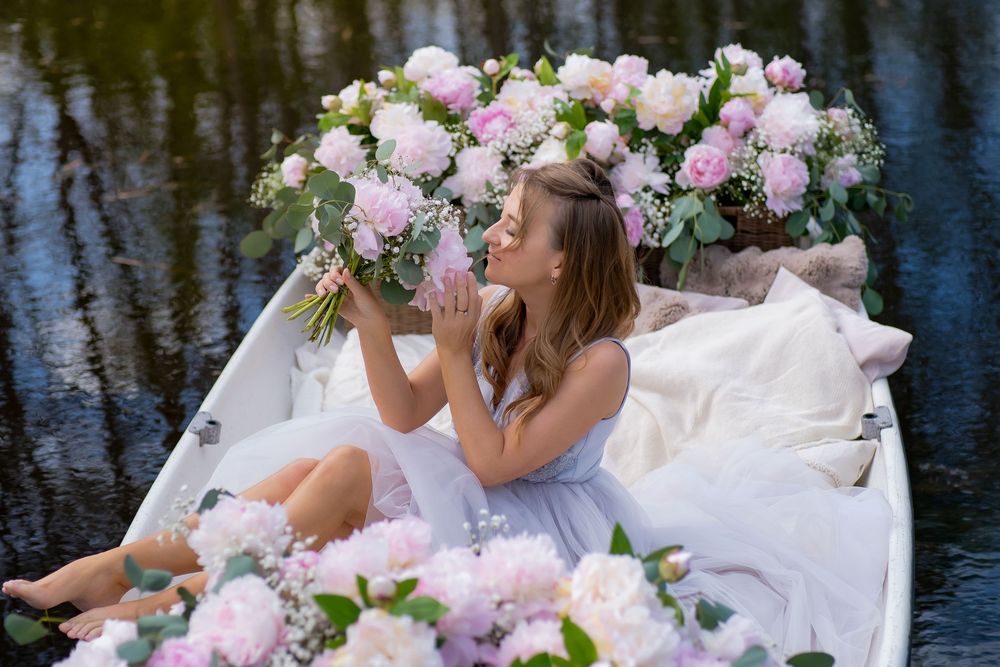 Romantic scene with soft pink peonies and white flowers surrounding someone in a lavender dress by the water's edge.
