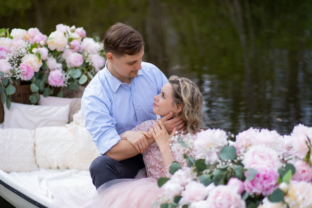 A romantic couple sits in a boat surrounded by pink and white floral arrangements on a peaceful lake.