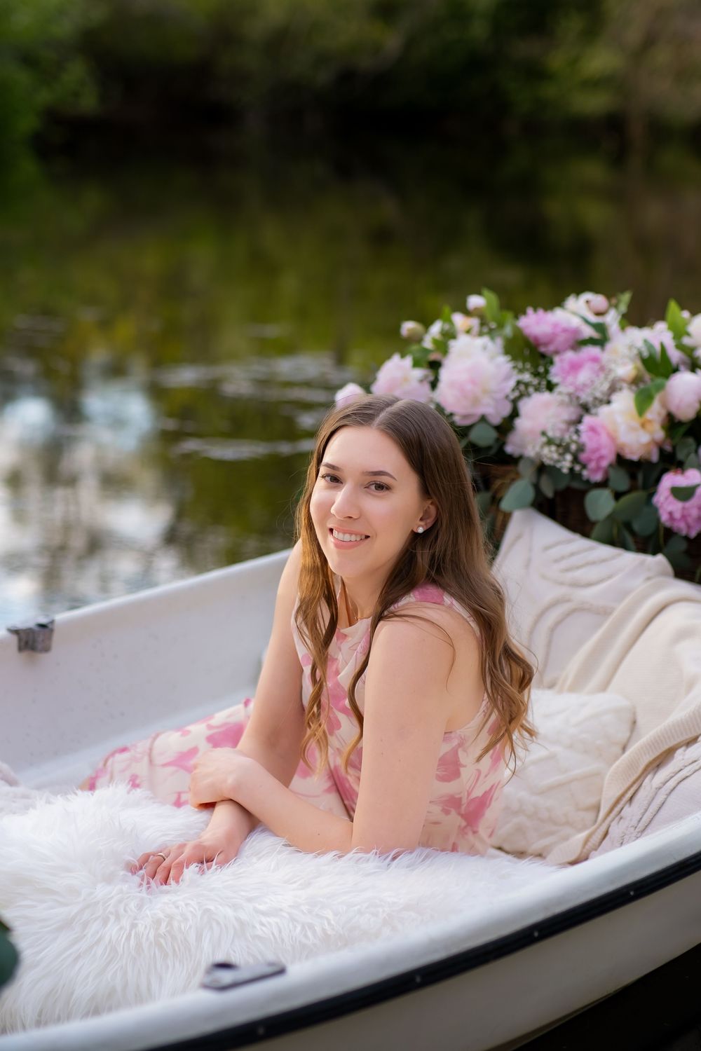 Someone poses gracefully on a white fur blanket by a lake with pink flowers in an outdoor portrait setting.