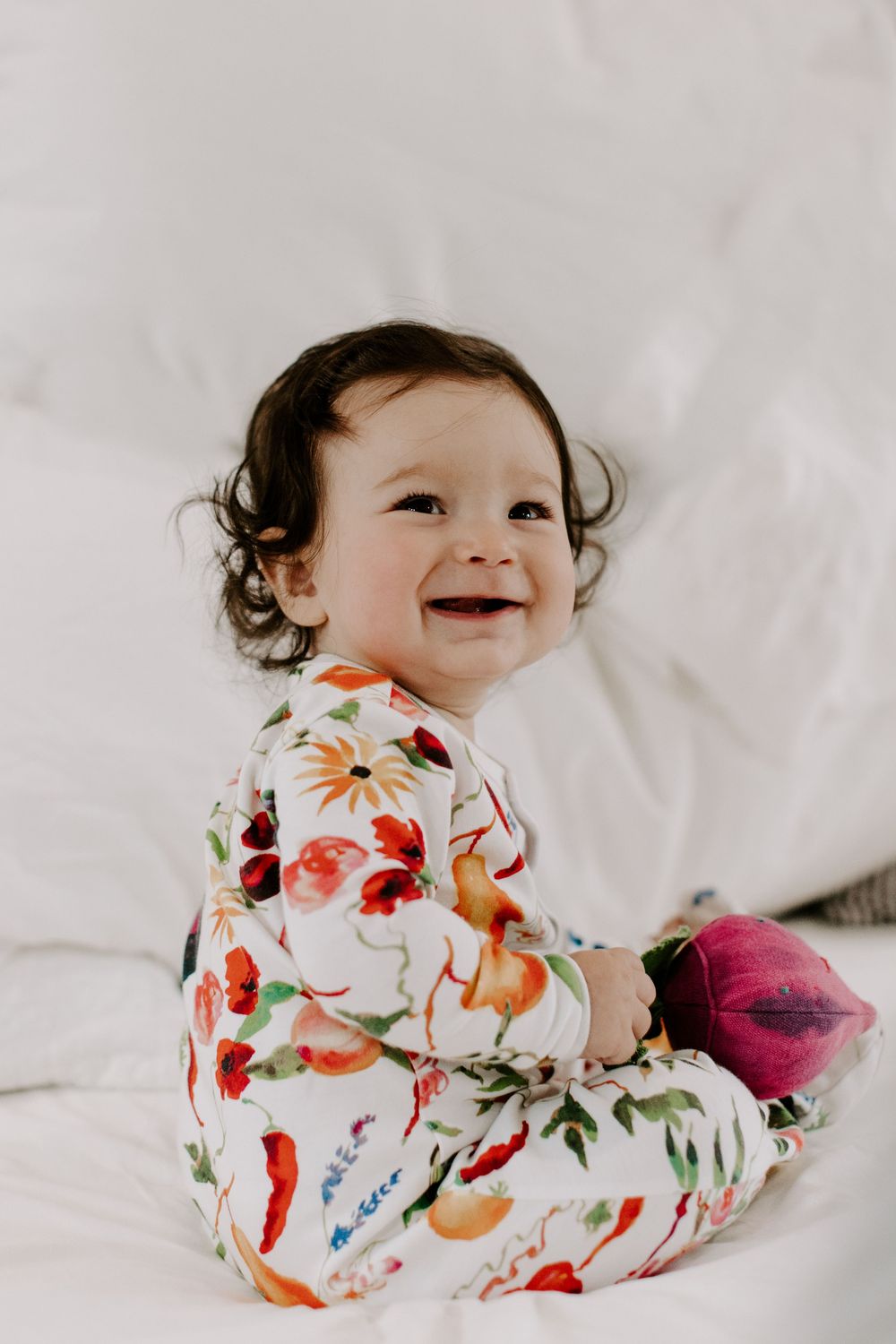 Smiling baby wearing colorful floral onesie lying on white bedding.