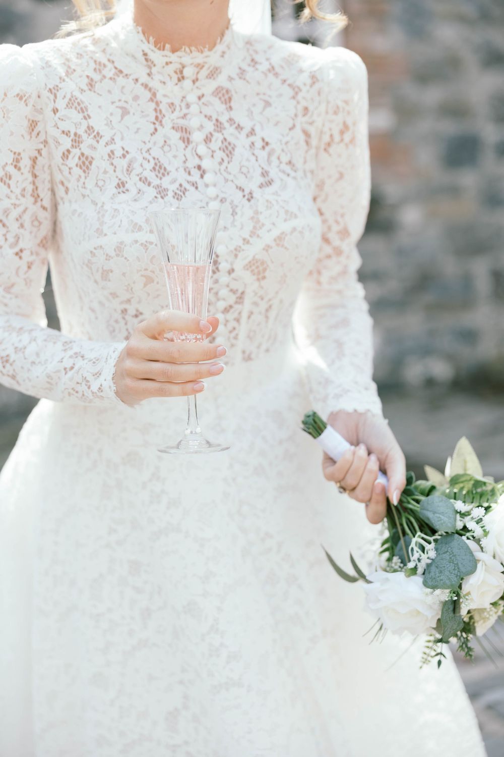 Close up of an elegant lace wedding dress with delicate details while holding a champagne glass and bouquet.