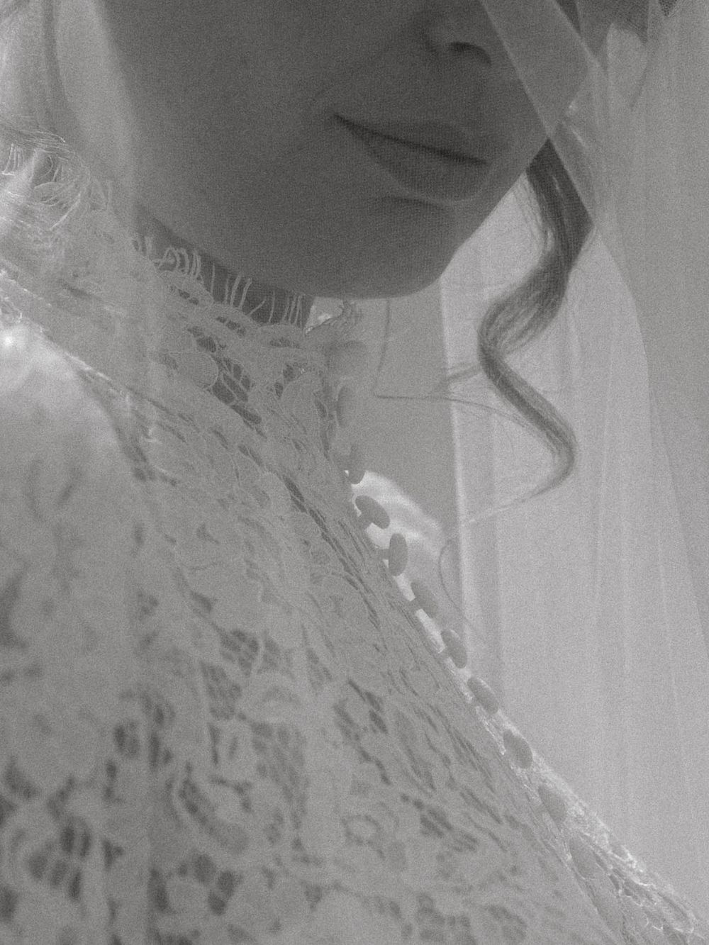 Black and white close-up detail of delicate white lace wedding dress with floral embroidery and soft curled hair strand.