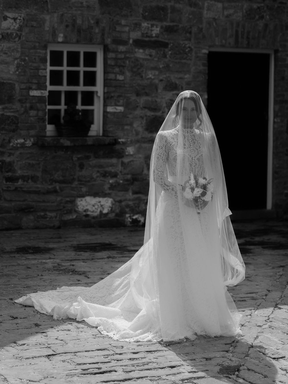 Elegant lace wedding gown and veil photographed from behind against a historic stone building.