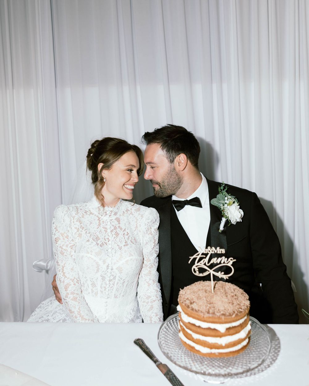 A romantic wedding couple shares a tender moment at their reception table with a rustic naked layer cake beside them.
