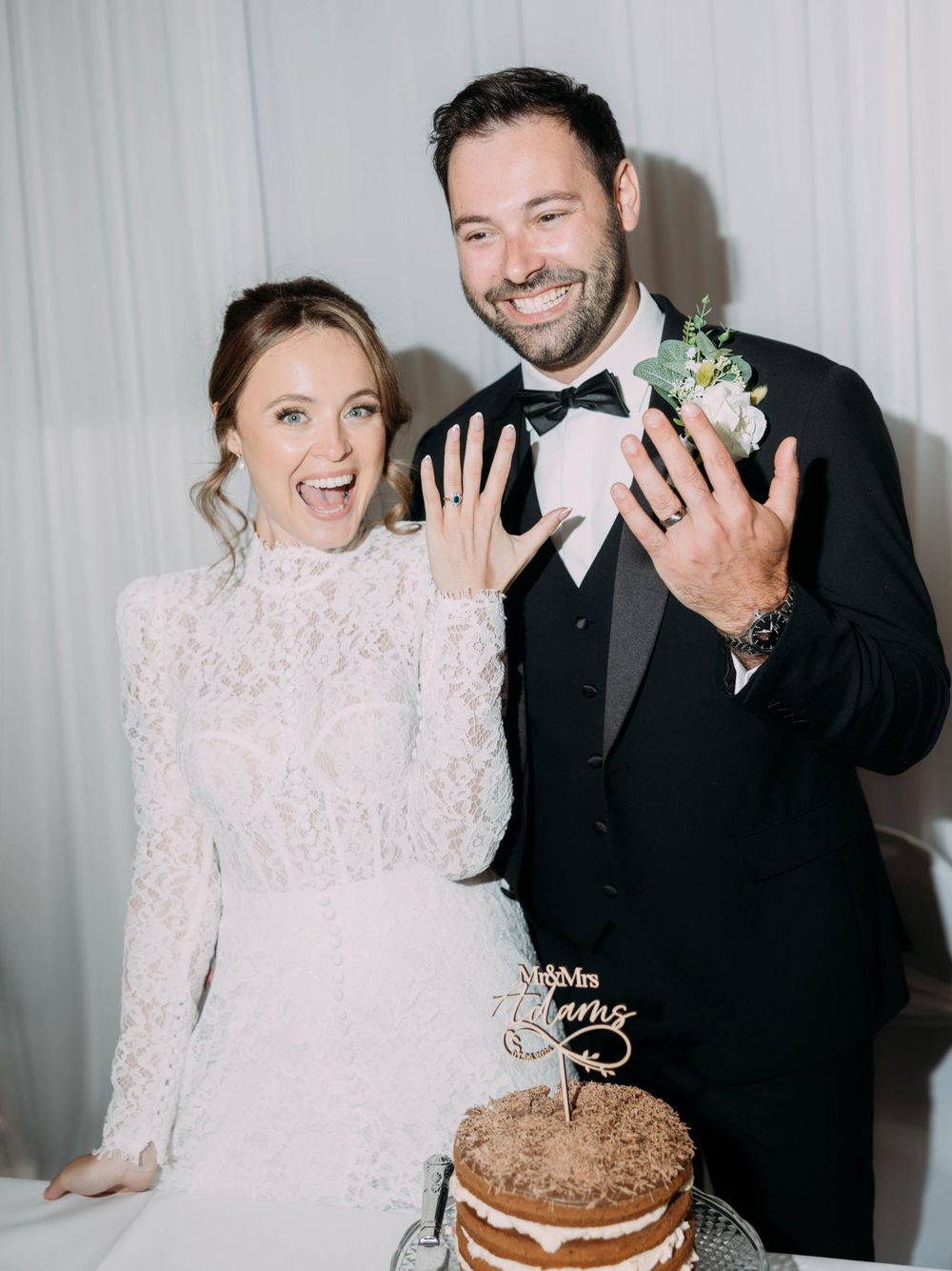 A joyful wedding couple showing off their rings beside their decorated chocolate cake at the reception.