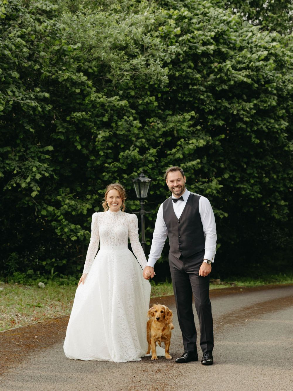 A couple in wedding attire holds hands while standing with their golden retriever dog on a tree-lined path.