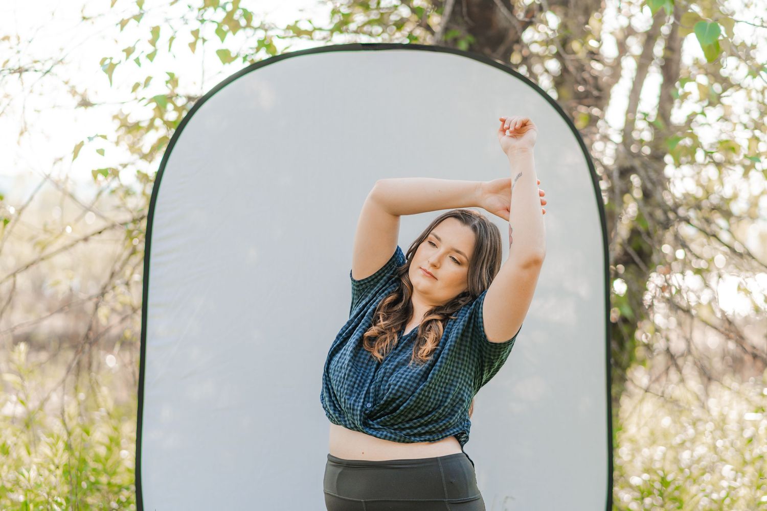 Woman posing with arms raised in front of a neutral pop-up backdrop outdoors, framed by trees and soft natural light in the PNW.