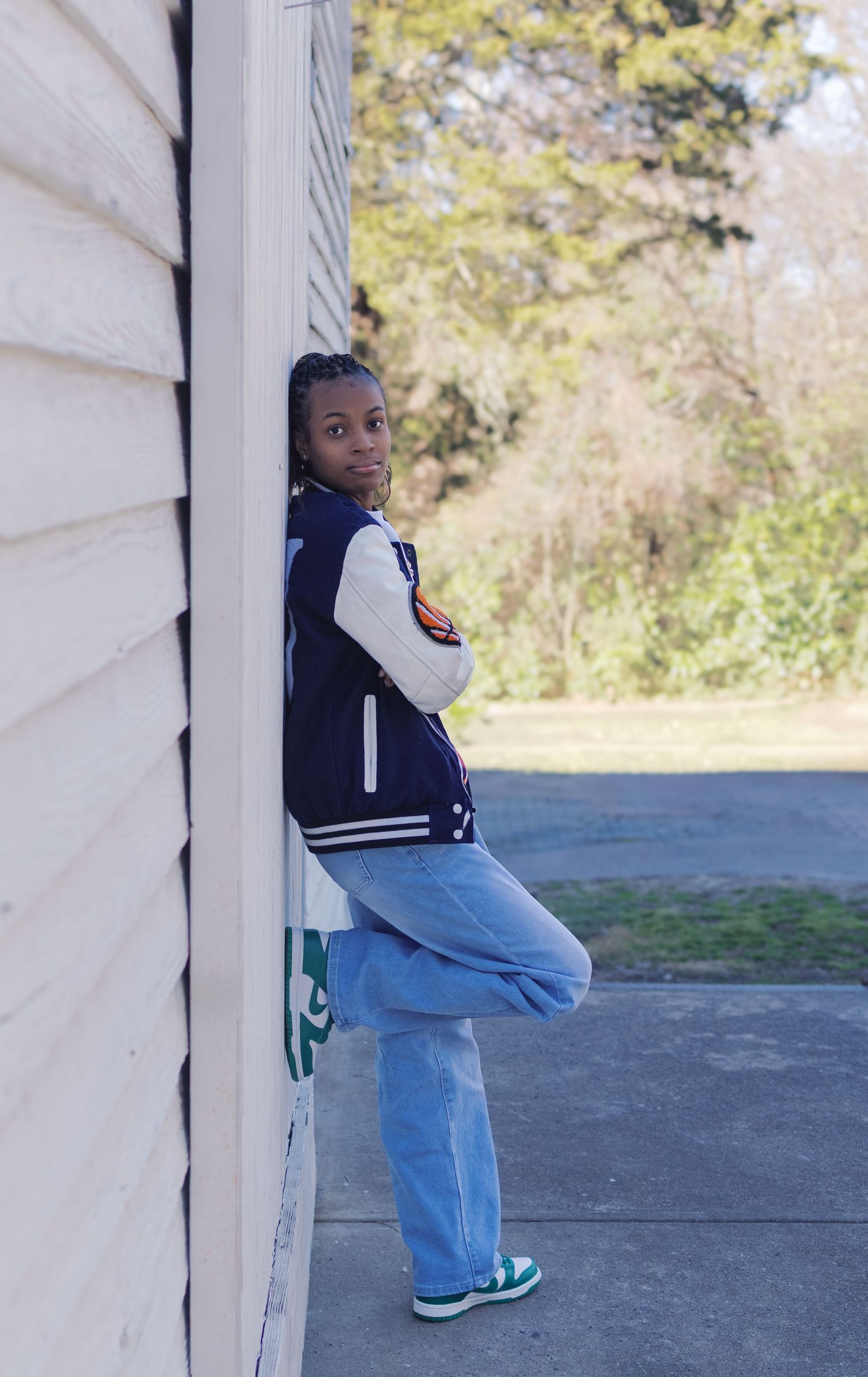 A casual portrait against white siding wearing a varsity jacket and jeans.