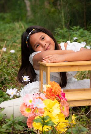 Young girl in white dress leaning on wooden stool surrounded by colorful flowers in garden.