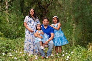 Family portrait with pregnant mother, father, and two daughters in blue outfits in green nature setting.