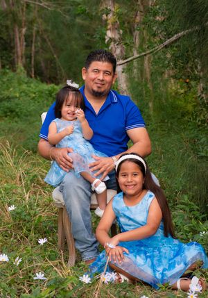 Father sitting outdoors holding toddler daughter while older daughter poses beside them in blue dresses.