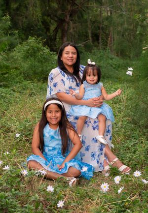 Mother in floral dress sitting outdoors with two daughters in matching blue outfits in green field.