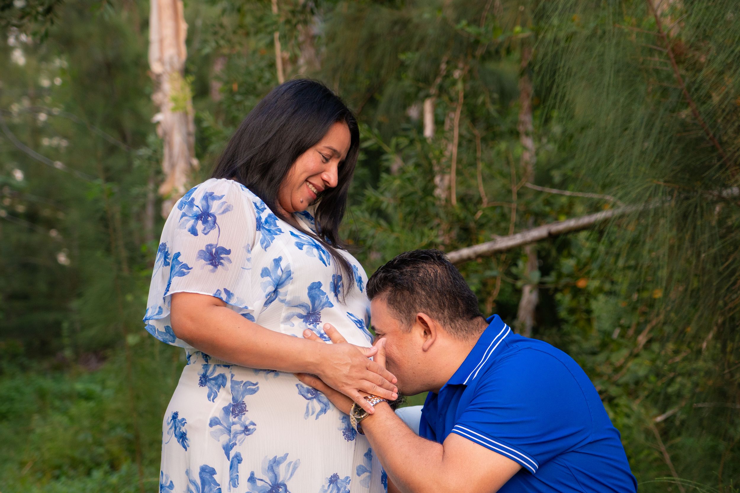 Pregnant woman in floral dress with partner kneeling and kissing her baby bump outdoors.