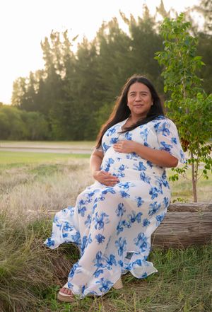 Pregnant woman in white and blue floral dress sitting outdoors in golden hour sunlight near trees.
