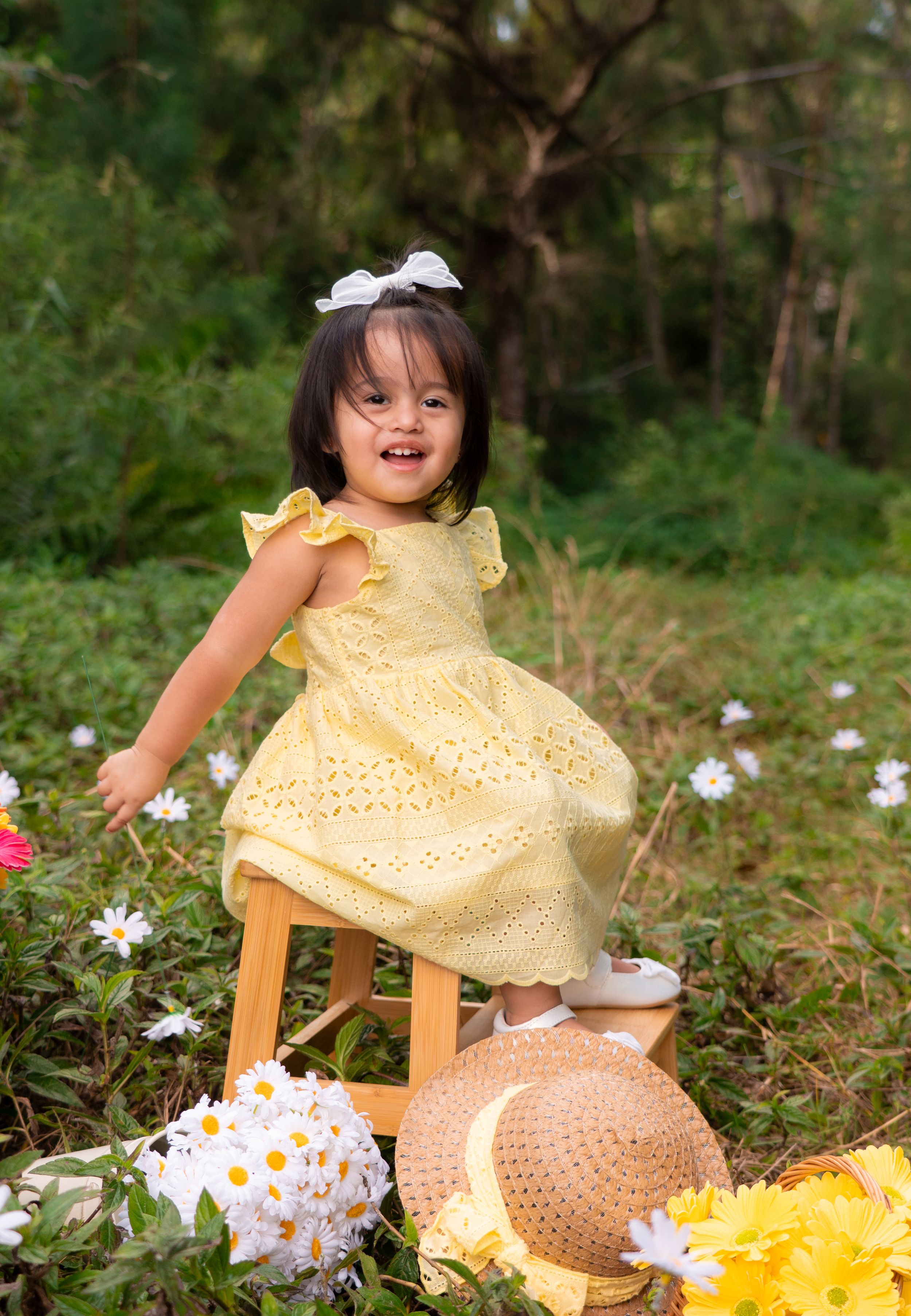 Happy toddler girl in yellow eyelet dress standing on wooden stool among white flowers outdoors.