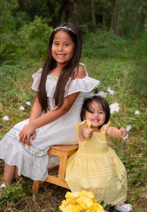 Two sisters posing outdoors, older girl in white dress seated with smiling toddler in yellow dress.