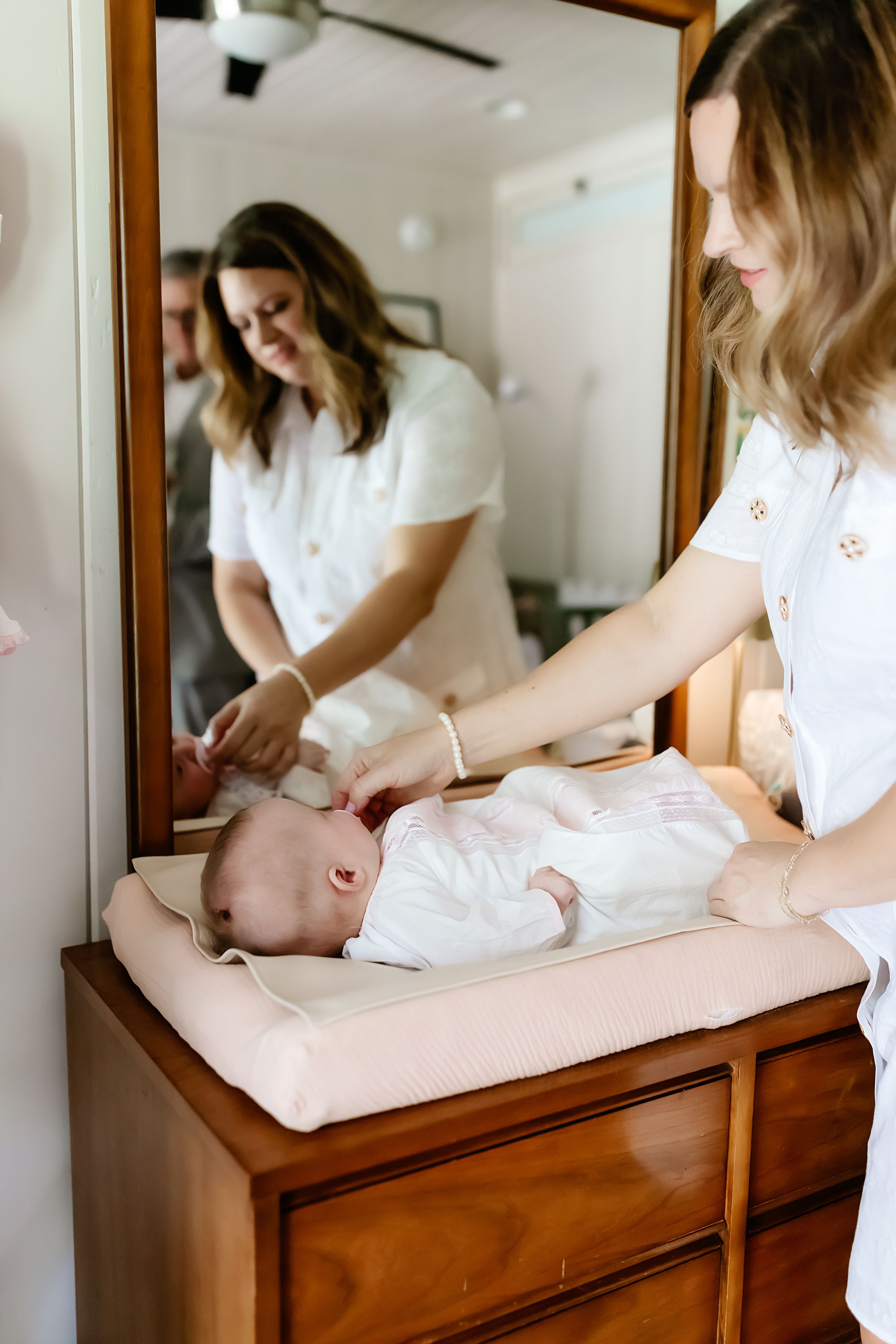 A baby gets changed on a wooden dresser while wearing white clothing.