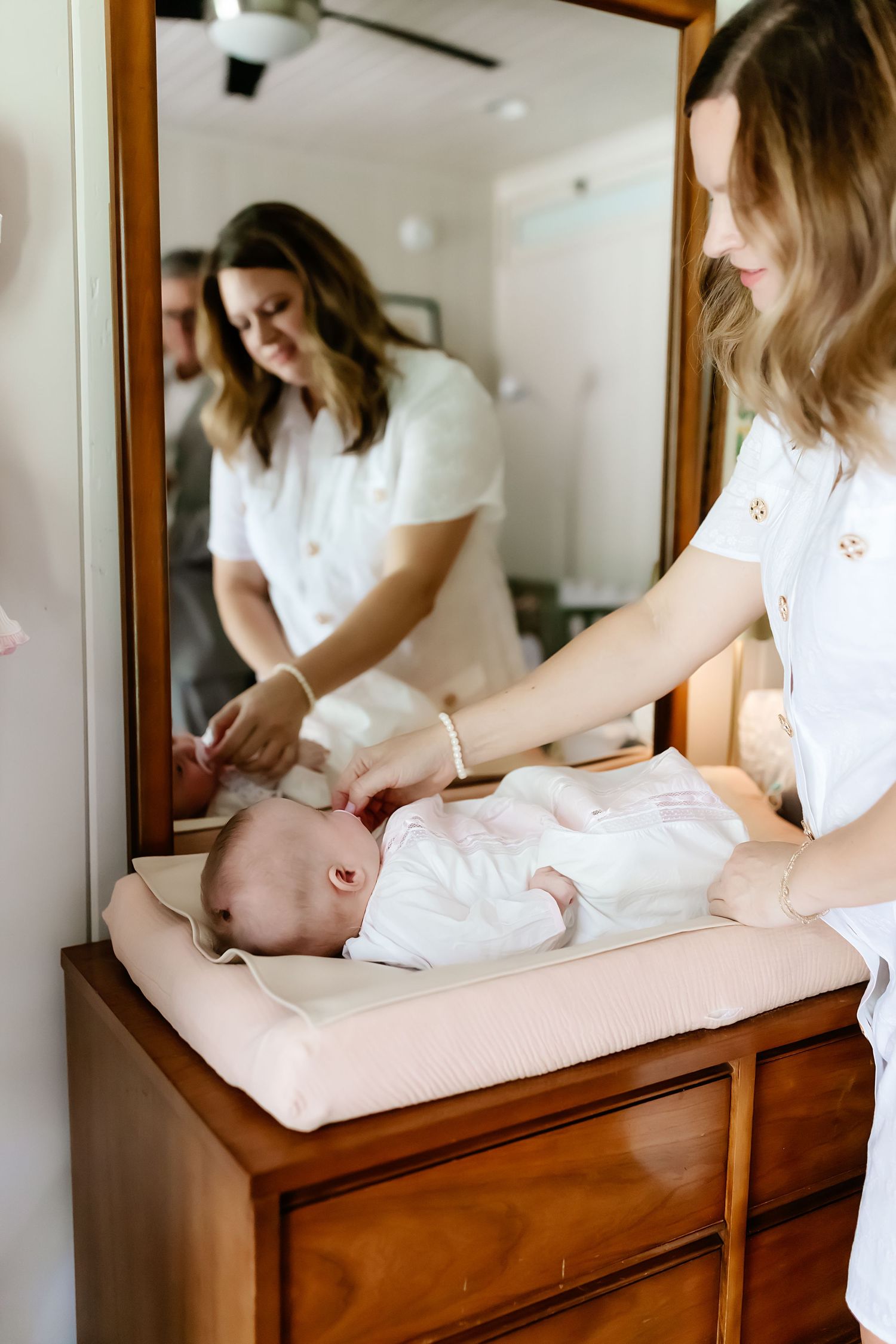 A baby gets changed on a wooden dresser while wearing white clothing.