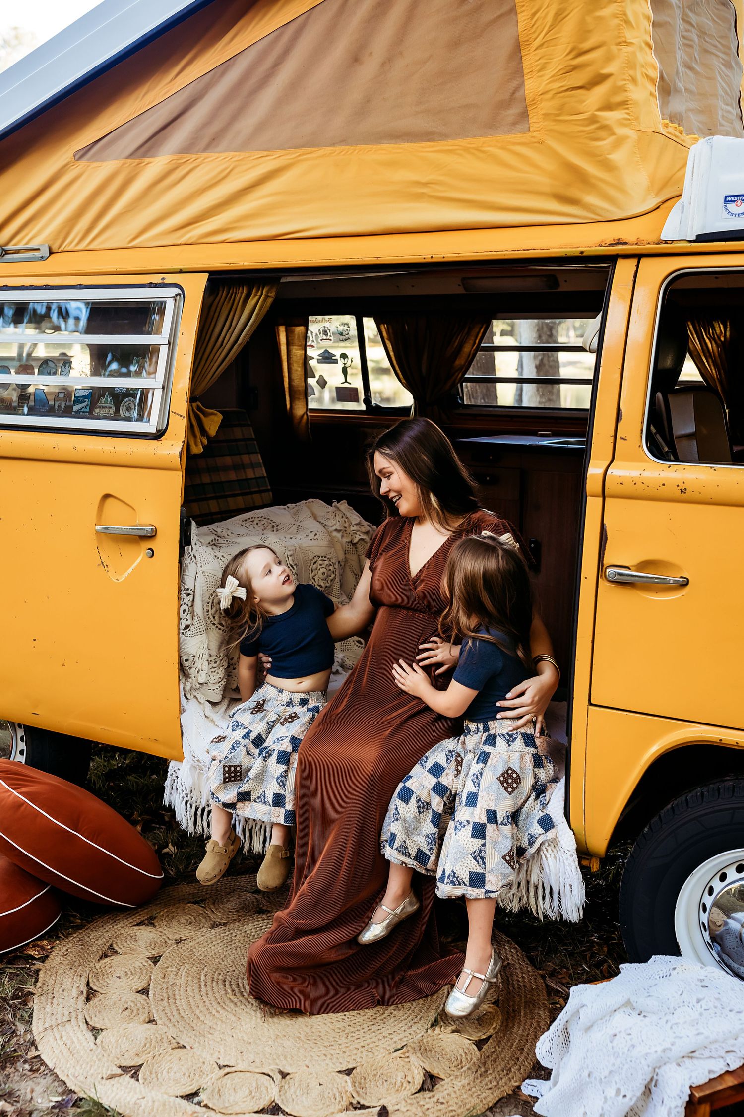 A vintage yellow Volkswagen van with pop-up camper top and people sitting inside on blankets and pillows.
