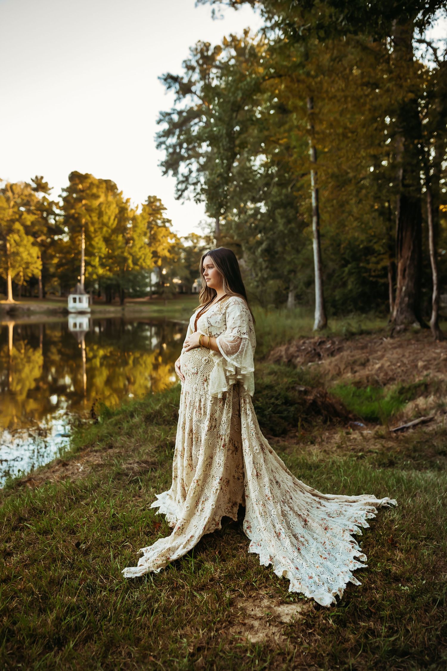 A person in a flowing lace dress stands by a serene lake at sunset with autumn trees reflecting in the still water.
