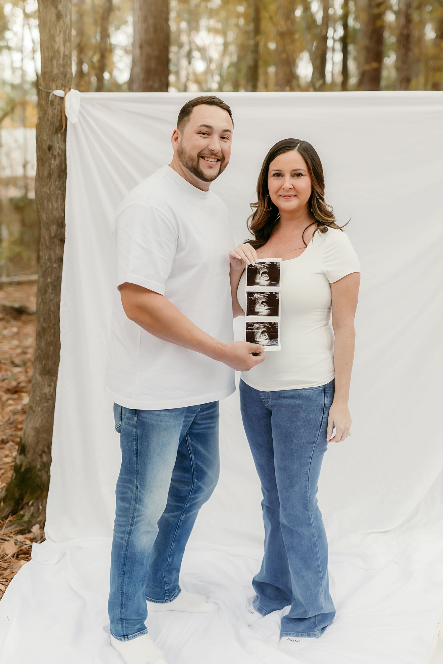 A couple stands against a white backdrop in a forest setting holding ultrasound photos to announce their pregnancy.