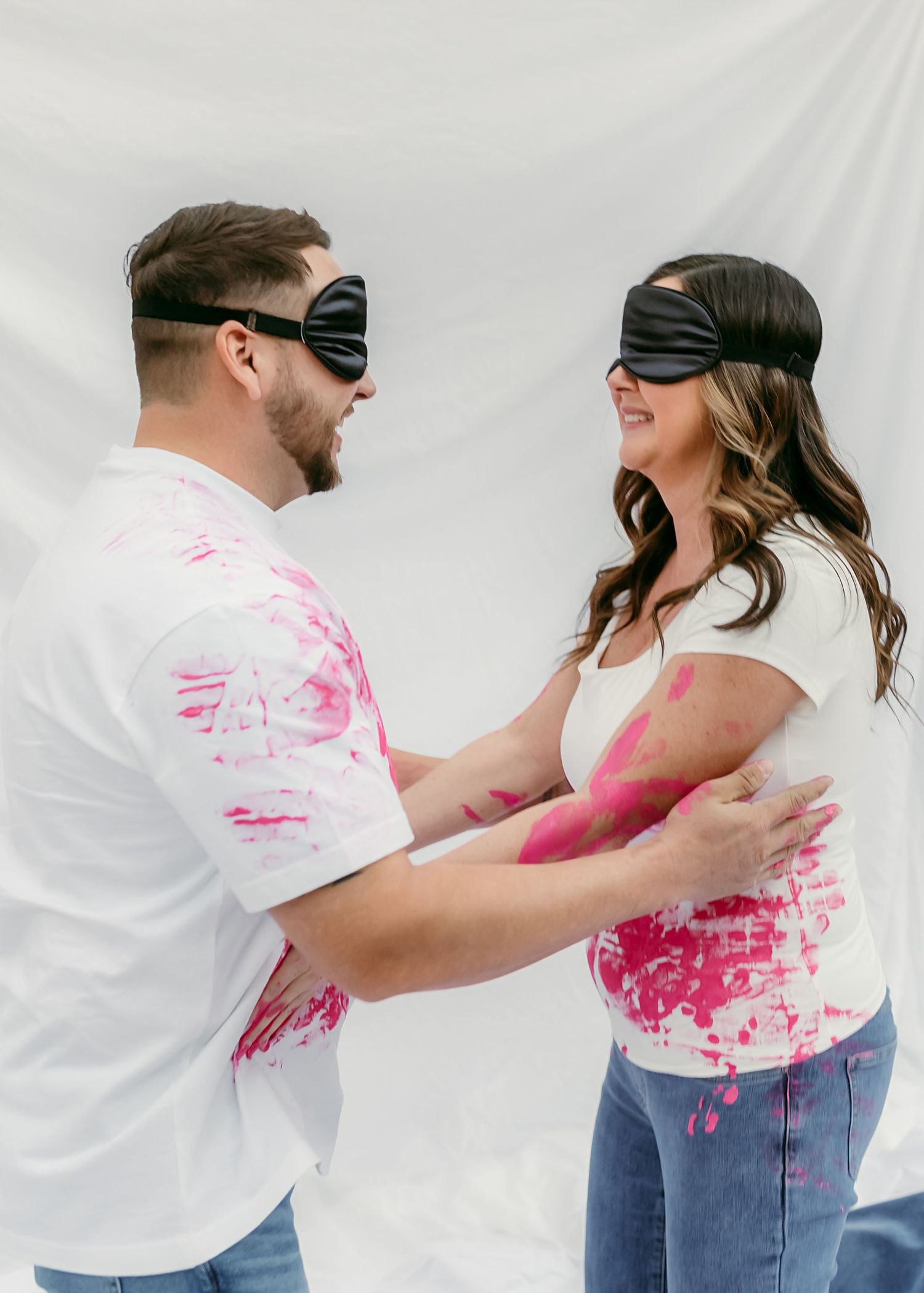 Two people wearing blindfolds wearing white t-shirts with pink paint stains laughing and embracing against a white backdrop.