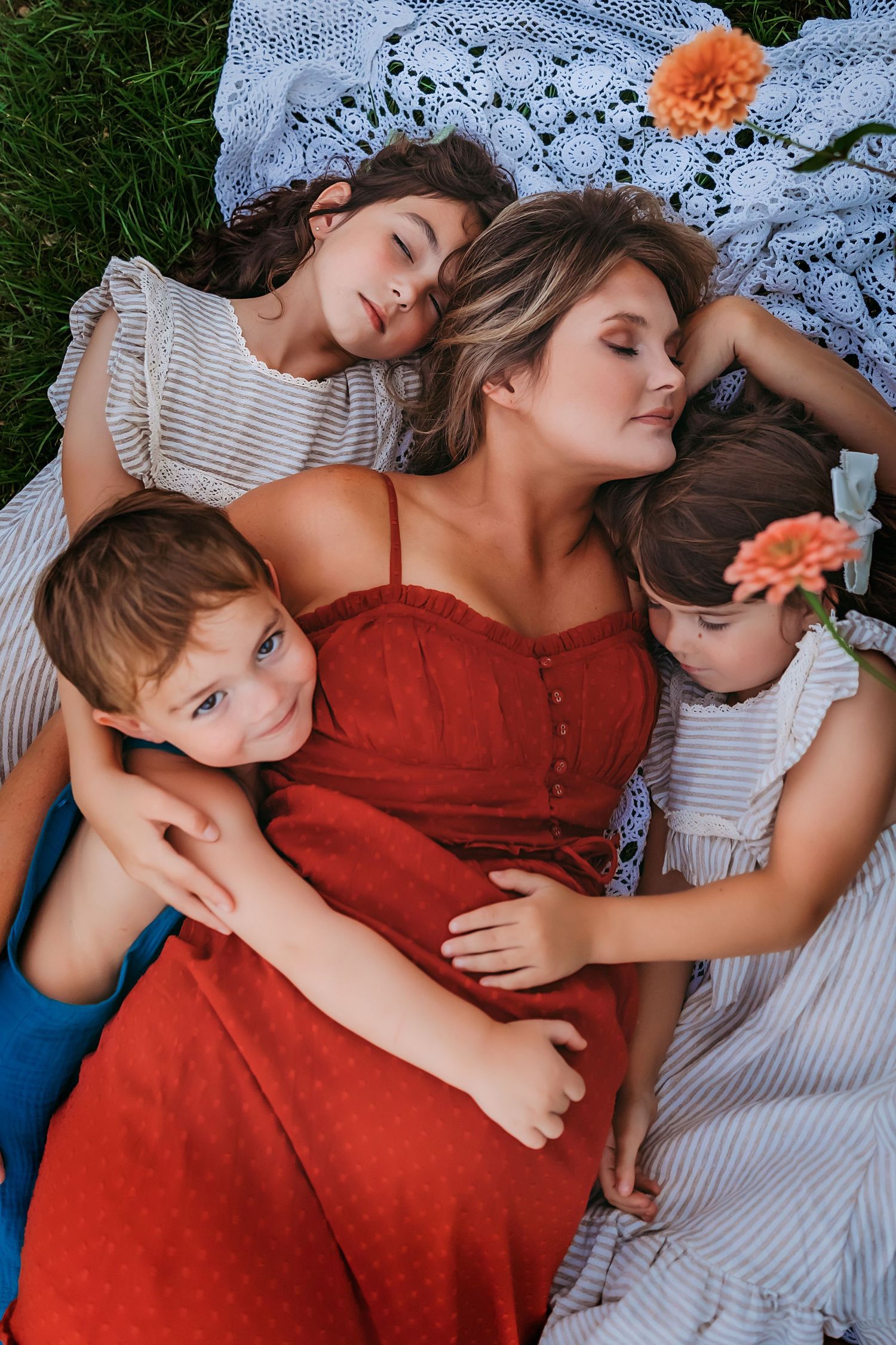 Family resting together on a white lace blanket in the grass with orange flowers and wearing red and striped clothing.