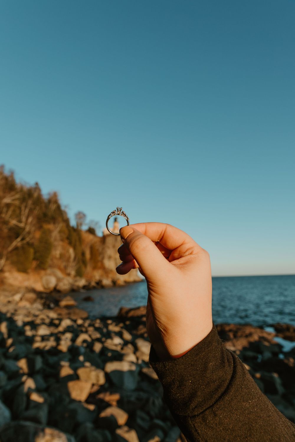 Hand holding a ring against a rocky coastline with blue water and cliffs during golden hour.