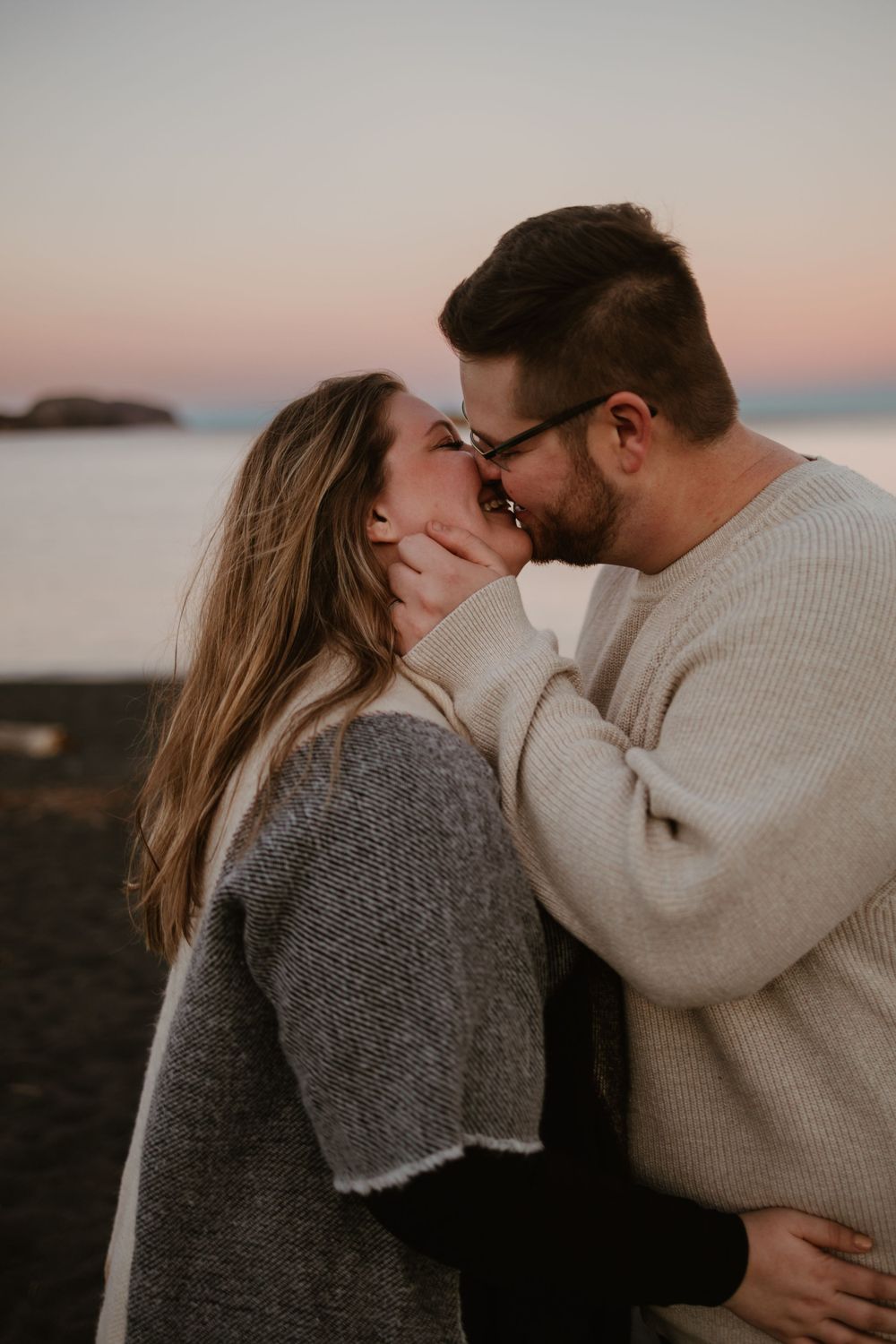 A romantic couple sharing a tender kiss at sunset by the coastline wearing cozy sweaters on a cool evening.