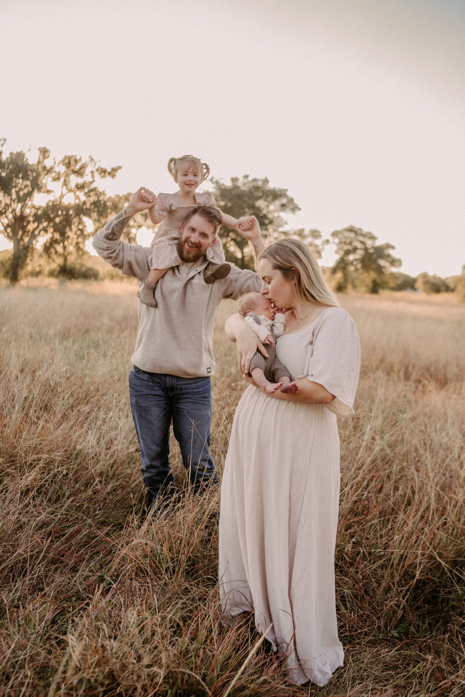 A family stands together in a golden field at sunset wearing neutral colored clothing in a dreamy outdoor setting.