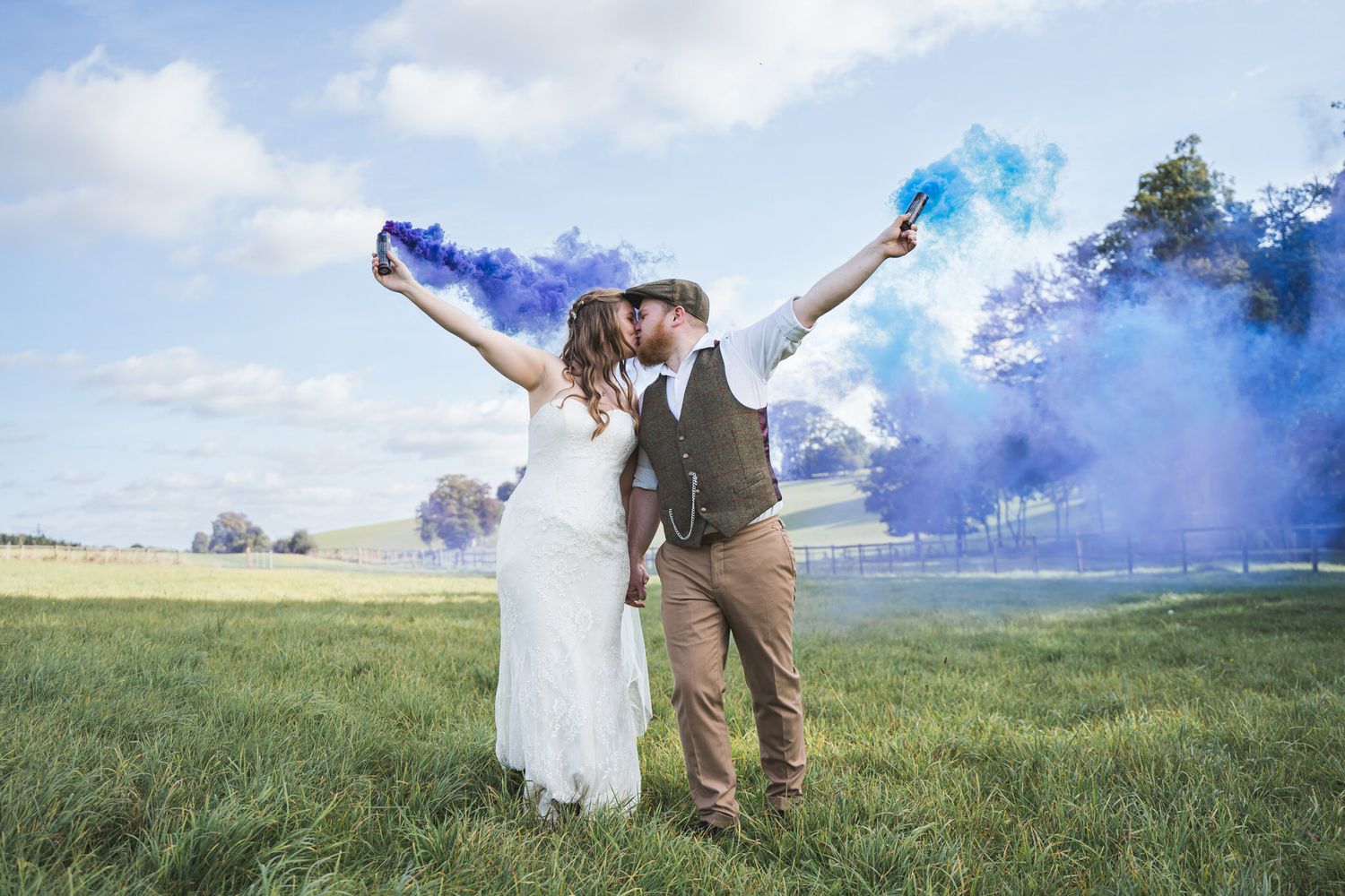 Bride and groom kissing in a field holding colourful blue and purple smoke bombs on their wedding day Herons Farm Wedding Venue
