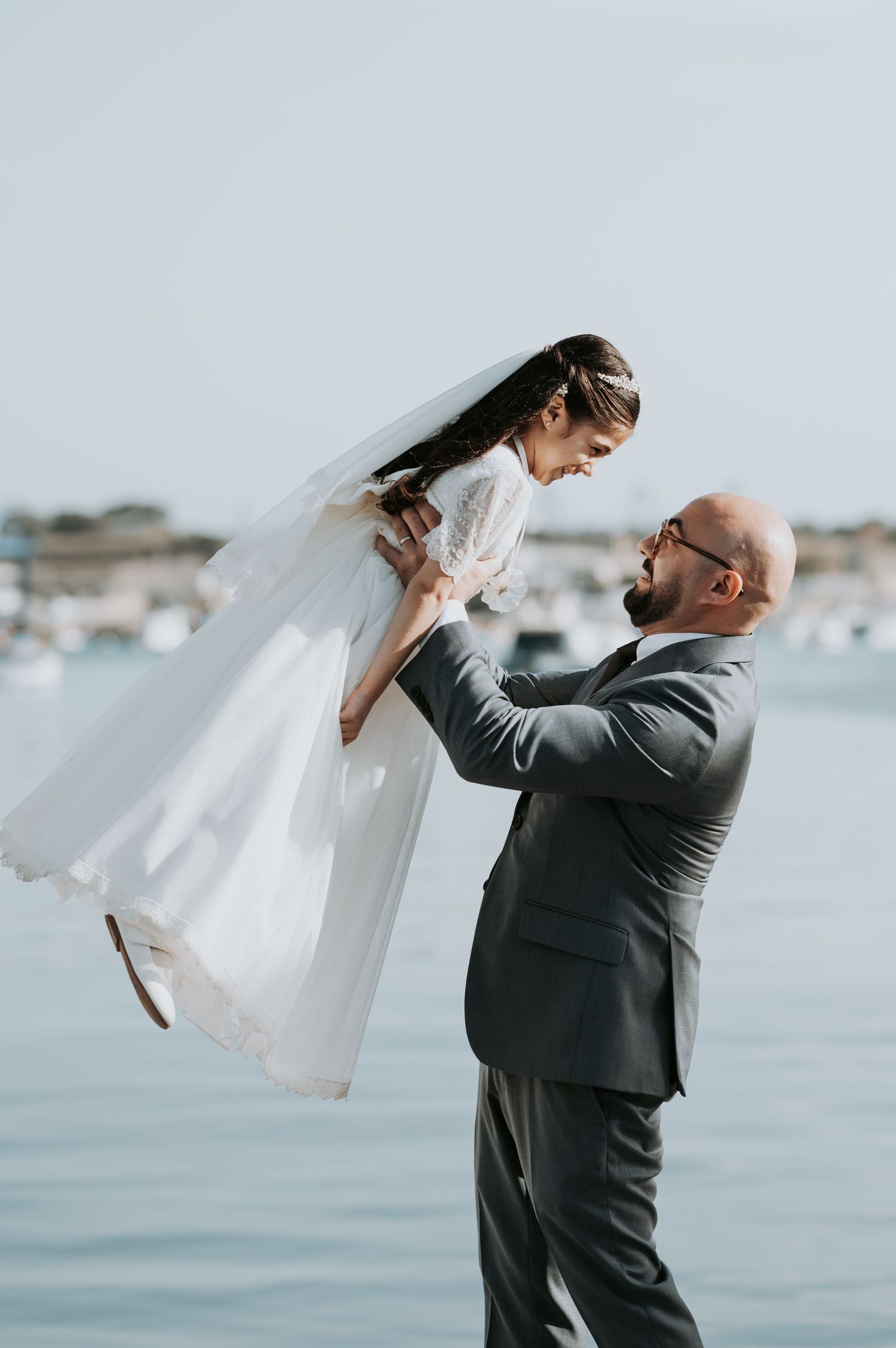 A touching moment between a bride in a flowing white dress and a man in a suit lifting her by the ocean.