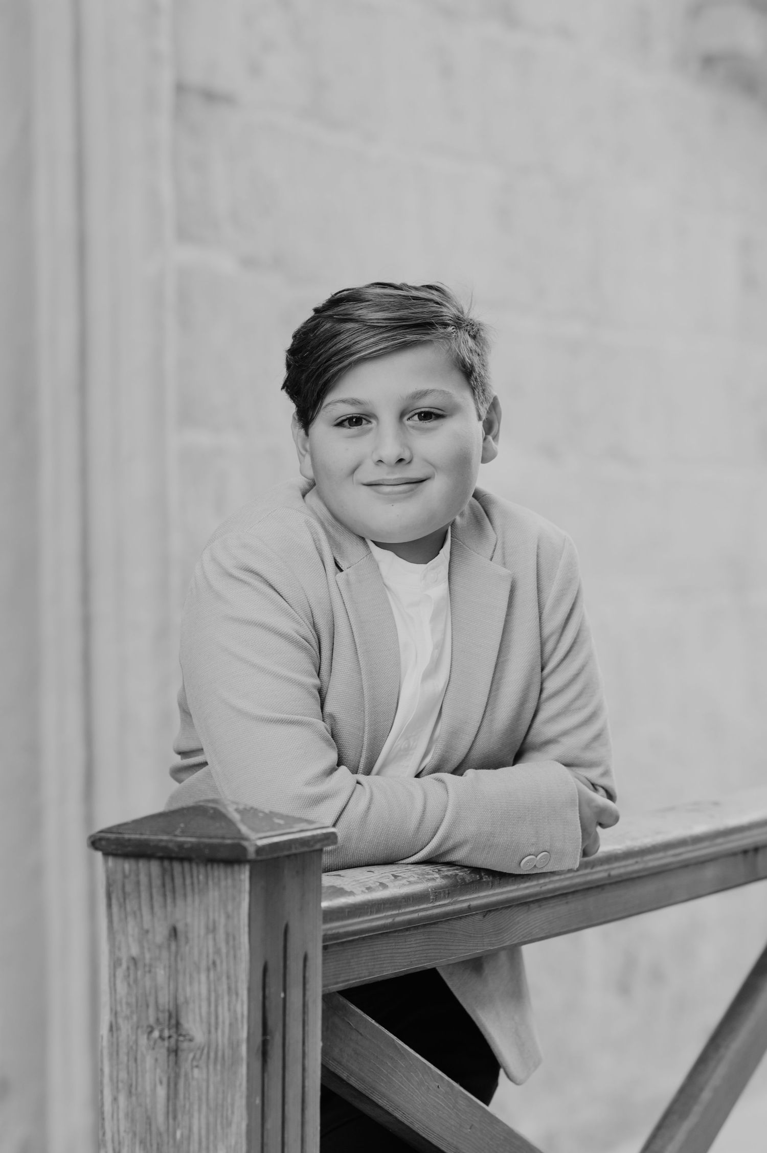 A person in a beige blazer and white shirt poses casually while leaning on a wooden surface.