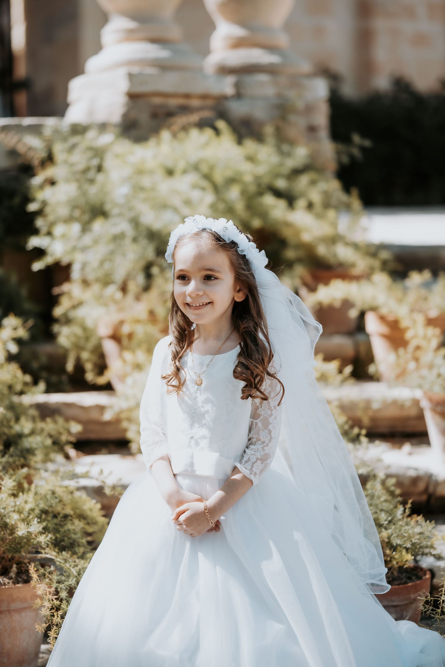 Child in white lace dress and veil twirling in garden setting with blooming flowers.