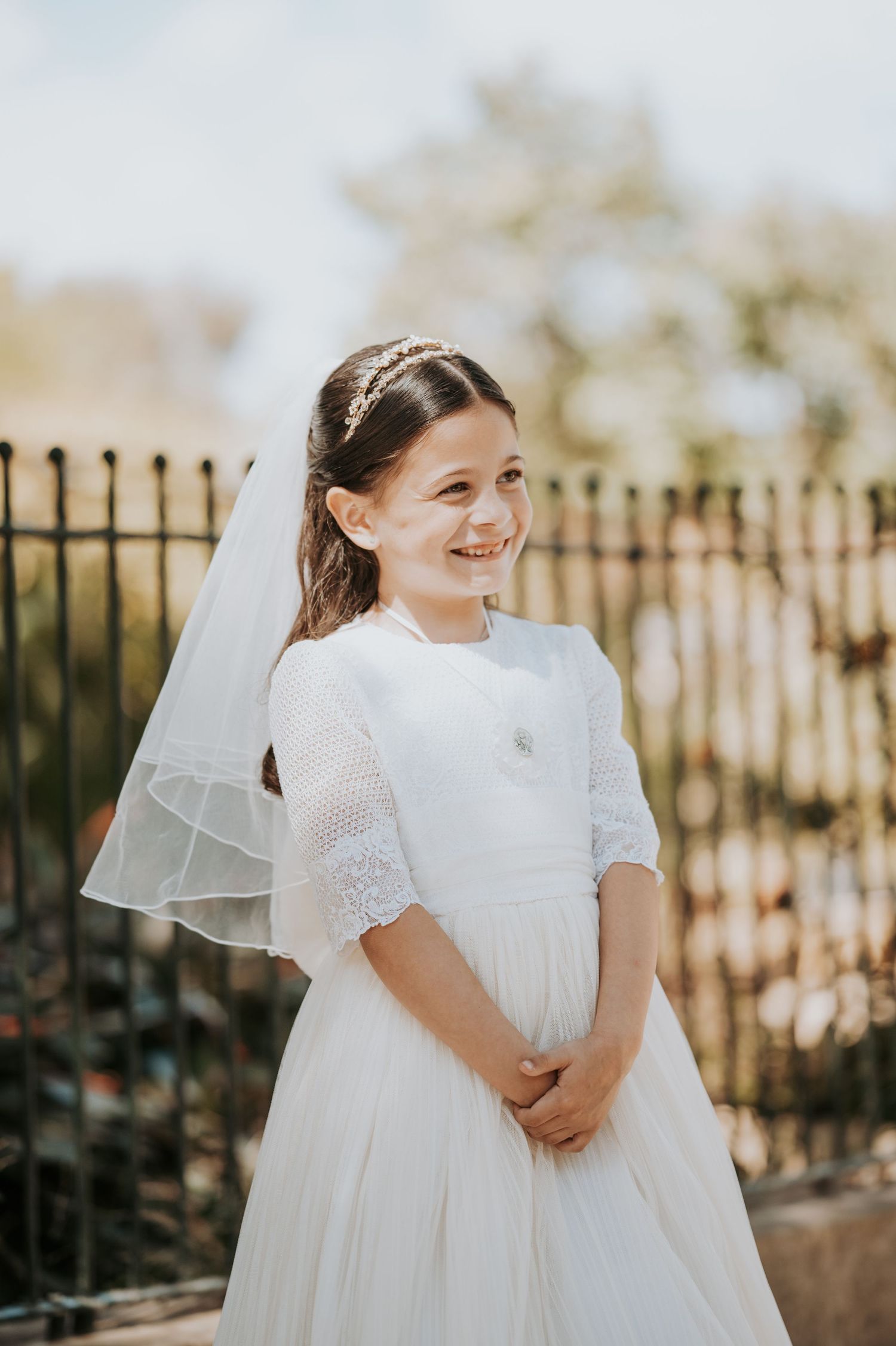 Young attendant in white lace dress with short puff sleeves and veil smiling in outdoor garden setting.