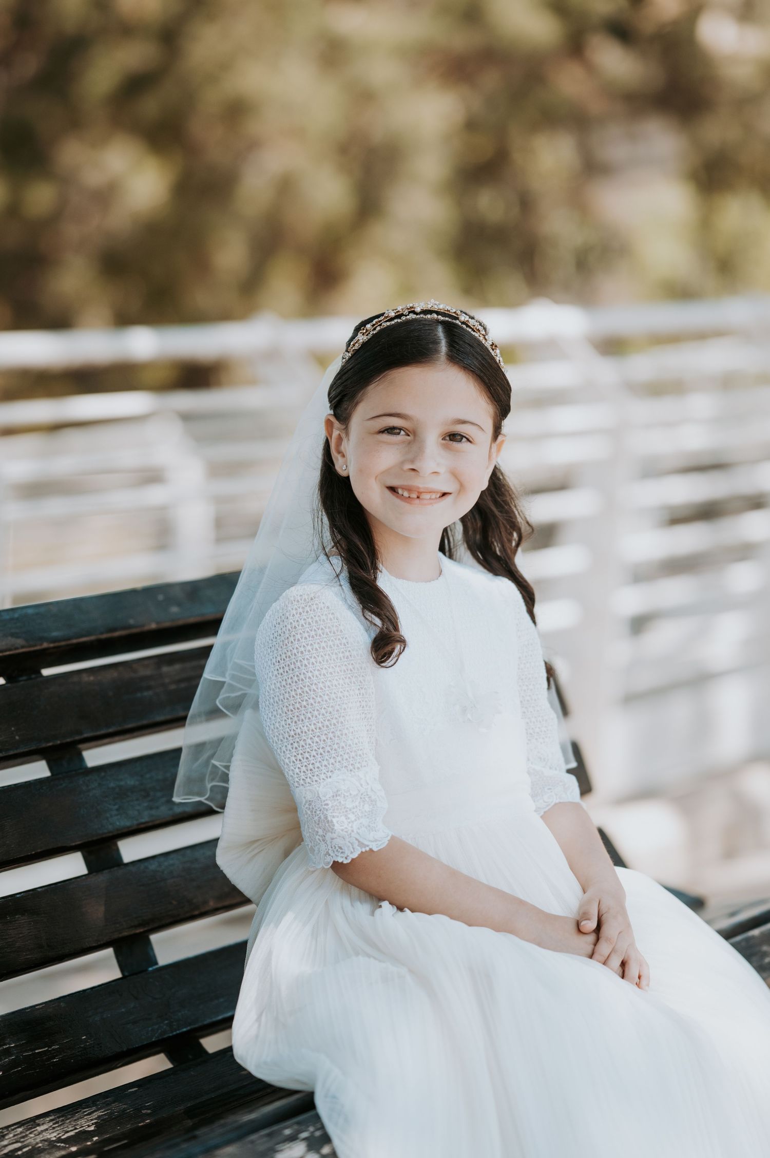 A child in a white dress sits on a wooden bench in natural outdoor lighting.