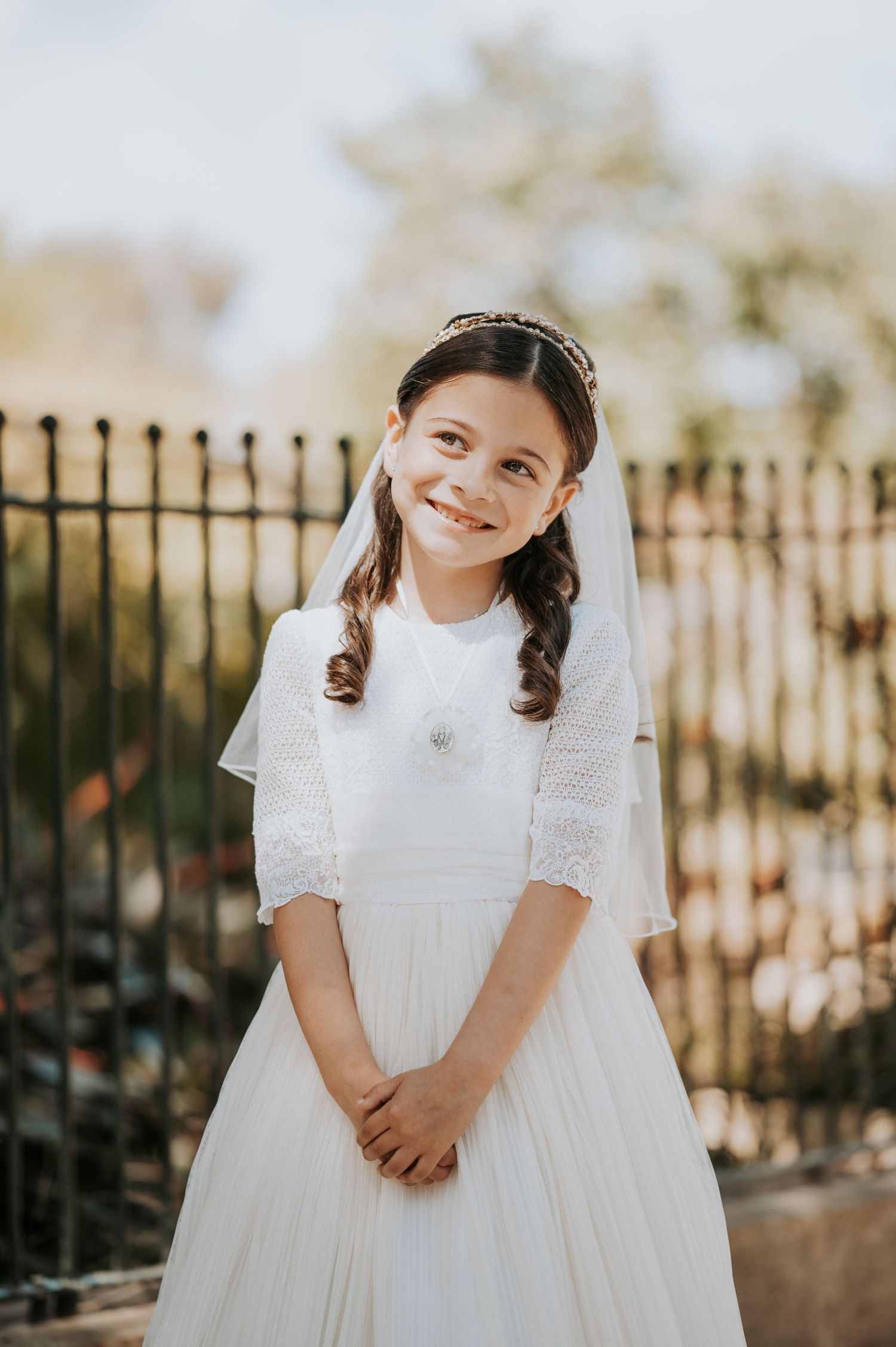 Child wearing elegant white communion dress posing against decorative iron fence in natural lighting.