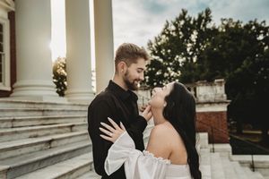 Romantic couple in elegant wedding attire embracing on grand steps with white columns and lush trees in background.