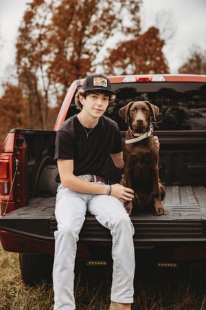 A person sits in a black pickup truck bed with a brown dog.