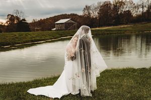 Bride in white gown with floral veil by a serene pond, rustic barn and autumn trees in background.