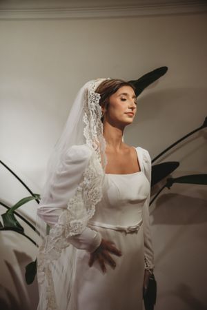 Bride in minimalist white wedding dress and lace veil poses elegantly in soft indoor lighting.