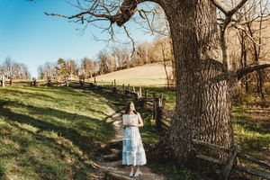 Young woman in blue dress standing beside a large tree on a sunny rural farm with wooden fence.