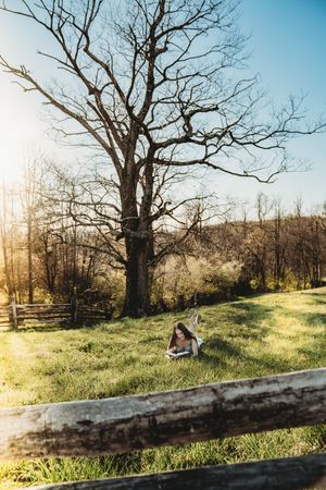 Young woman reading a book on green grass beneath a bare tree in a sunny rural landscape with wooden fence.
