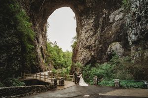Bride and groom embrace under a massive natural rock arch surrounded by lush greenery at a cave wedding venue.