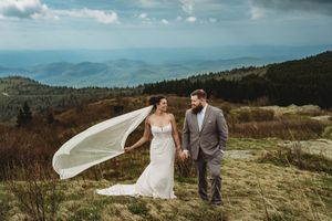 Bride and groom hold hands on a mountain summit, her veil flowing in the wind with scenic valley views behind them.