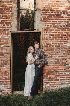A romantic couple poses against a rustic brick wall with red flowers in the background.