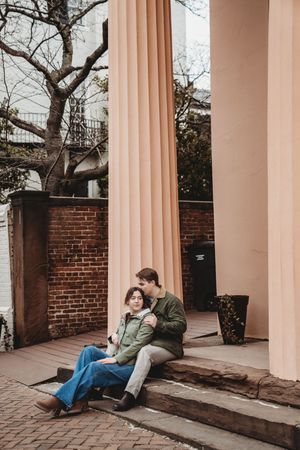 A couple sits together on brick steps beside a pink column and brick wall on an autumn day.